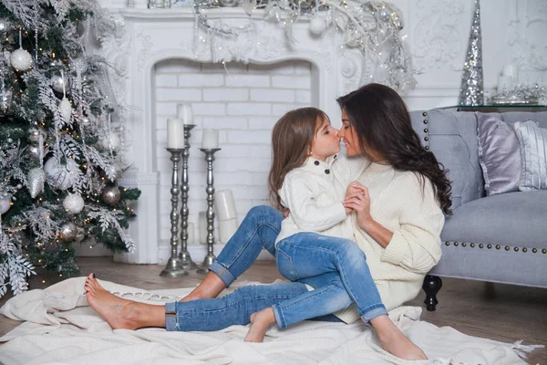 Yeni yıl arifesinde mutlu bir aile. Süslü süveter içinde güzel anne ve tatlı kız eğleniyor ve süslü bir Noel ağacının altında sarılıyor. Şömineli ve gri kanepeli ev klasiği..