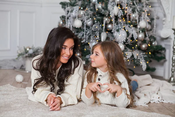 Happy family on New Year Eve. Young beautiful mother and little cute daughter in white cozy sweaters and blanket have fun and hug under a decorated Christmas tree. Festive home classic interior.