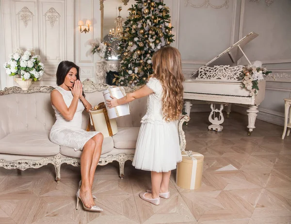 Mutlu Noeller ve mutlu tatiller. Neşeli anne ve tatlı kızı, beyaz klasik iç mekanda bir piyanonun arka planına ve süslü bir Noel ağacına karşı hediyelerini değiş tokuş ediyorlar. Yeni Yıl 2021 