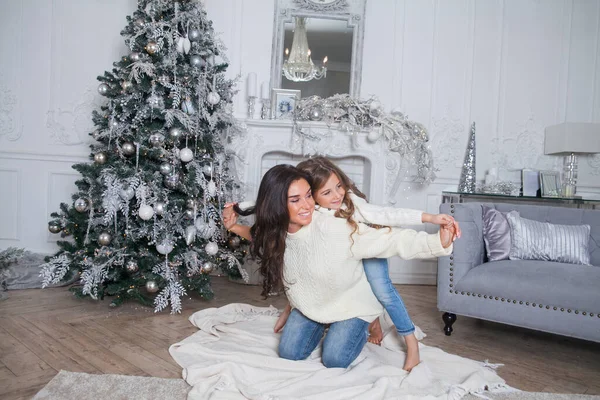 Happy family on New Year Eve. Beautiful mother and cute daughter in white cozy sweaters have fun and hug under a decorated Christmas tree. Festive home classic interior with fireplace and grey sofa.