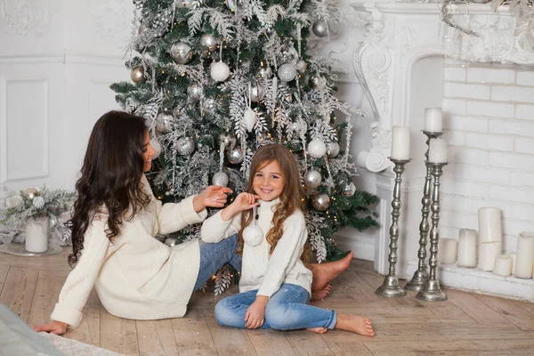 Happy family on New Year Eve. Young beautiful mother and little cute daughter in white cozy sweaters and blanket have fun and hug under a decorated Christmas tree. Festive home classic interior.