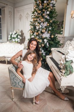 Mutlu Noeller ve mutlu tatiller. Neşeli anne ve beyaz klasiklerde beyaz bir piyanoda süslü bir Noel ağacının arka planında çalan tatlı kızı. Yeni Yıl 2021 