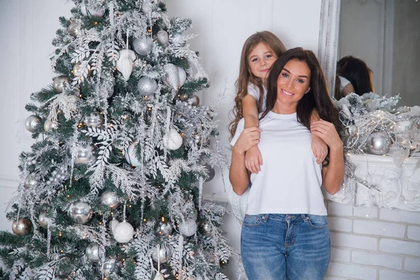 Happy family New Year Eve. Young beautiful mother and little cute daughter in white shirts and jeans having fun and hugging under a decorated Christmas tree. Festive home classic interior with mirror.