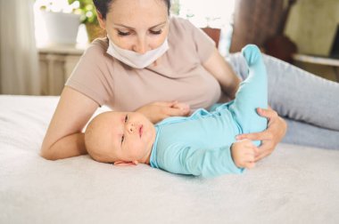 Koruyucu yüz maskesi takmış güzel bir anne kucağında yeni doğmuş oğlunu virüslerden ve enfeksiyonlardan koruyor. Coronavirus covid-19 önleme. Ev karantinası. Evde mutlu bir aile.