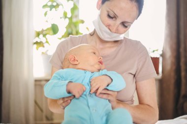 Koruyucu yüz maskesi takmış güzel bir anne kucağında yeni doğmuş oğlunu virüslerden ve enfeksiyonlardan koruyor. Coronavirus covid-19 önleme. Ev karantinası. Evde mutlu bir aile.