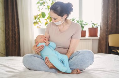 Koruyucu yüz maskesi takmış güzel bir anne kucağında yeni doğmuş oğlunu virüslerden ve enfeksiyonlardan koruyor. Coronavirus covid-19 önleme. Ev karantinası. Evde mutlu bir aile.