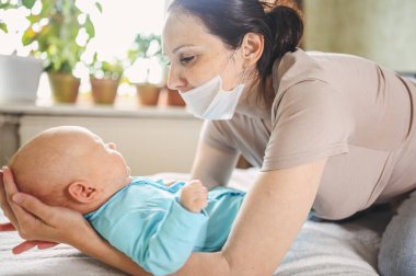 Koruyucu yüz maskesi takmış güzel bir anne kucağında yeni doğmuş oğlunu virüslerden ve enfeksiyonlardan koruyor. Coronavirus covid-19 önleme. Ev karantinası. Evde mutlu bir aile.