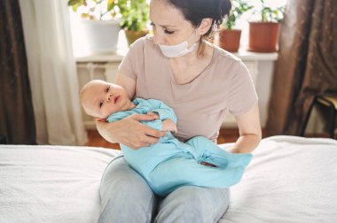 Koruyucu yüz maskesi takmış güzel bir anne kucağında yeni doğmuş oğlunu virüslerden ve enfeksiyonlardan koruyor. Coronavirus covid-19 önleme. Ev karantinası. Evde mutlu bir aile.