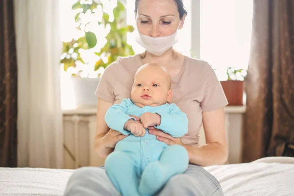 Beautiful mother in protective face mask holding her little cute newborn son protecting him from viruses and infections. Coronavirus covid-19 prevention. Home quarantine. Happy family at home.