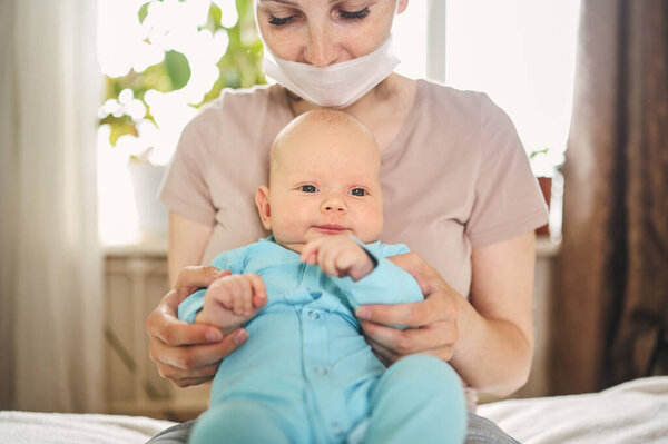 Красивая мать в защитной маске держит своего маленького милого новорожденного сына, защищающего его от вирусов и инфекций. Коронавирусный ковид-19 профилактика. Домашний карантин. Счастливая семья дома.