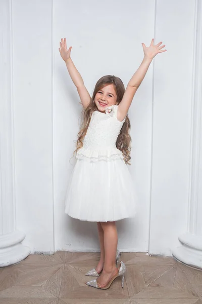 Little princess - excited emotional cute girl in fashion white dress having fun and wearing big mothers sparkle high heels shoes on white background. Free space for text mockup