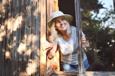 Mutlu, güler yüzlü, yaşlı bir kadın, saman şapkalı, eski ahşap köy evinde, pencerenin önünde poz verirken eğleniyor. Emekli yaşlılar konsepti. Kır evinde karantina