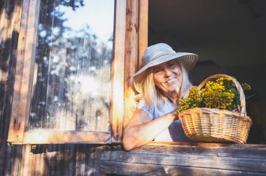Mutlu gülümseyen yaşlı kadın kırsal kesimdeki ahşap köy evinde çiçek sepetli, açık pencerede poz verirken eğleniyor. Emekli yaşlılar konsepti. Kır evinde karantina