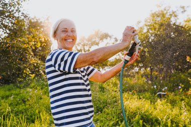 Mutlu yaşlı bir kadın yaz bahçesinde çiçekleri hortumla sularken eğleniyor. Arka planda bir damla su. Çiftçilik, bahçıvanlık, tarım, yaşlı insanlar. Çiftlikte organik sebzeler yetiştiriyor.. 