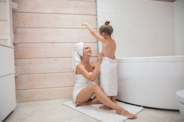 Mutlu sevgi dolu aile birlikte vakit geçiriyor. Anne ve kızı beyaz banyoda öpüşüp sarılıyorlar. Kafalarında ve vücutlarında havlular olan kadın ve kız. 