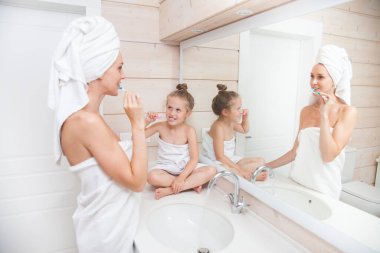 Dişlerini fırçalayan ve birlikte vakit geçiren mutlu bir aile. Anne ve kızı beyaz banyoda öpüşüp sarılıyorlar. Kafalarında ve vücutlarında havlular olan kadın ve kız. 