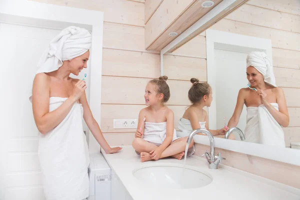 Happy loving family brushing teeth and spending time together. Mother and her daughter child girl kissing and hugging in white bathroom. Woman and girl with towels on their heads and bodies