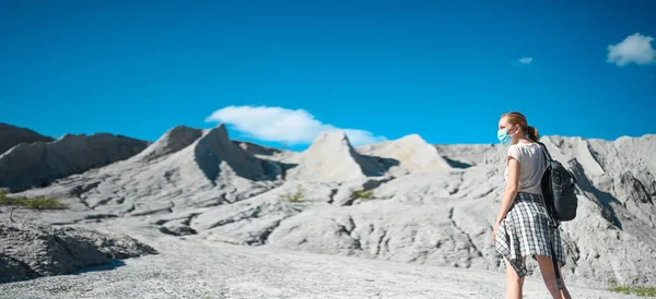 Genç sarışın kadın Covid 19 önleme için yüz koruyucu maske takarak beyaz dağlara doğru yürüyor. Sırt çantalı genç gezgin öğrenci. Corona virüsü ve yerli turizm kavramı.