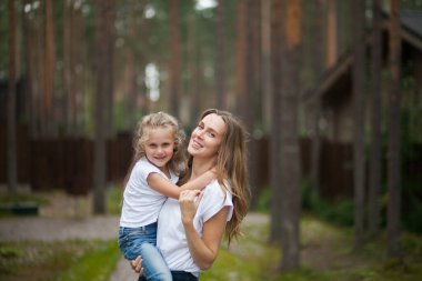 Mutlu gülümseyen annesi ve beyaz tişörtlü ve kot pantolonlu sevimli duygusal kızı kırsal alanda kucaklaşıp eğleniyorlar. Yazı ailemle geçiriyorum.