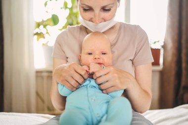 Koruyucu yüz maskesi takmış güzel bir anne kucağında yeni doğmuş oğlunu virüslerden ve enfeksiyonlardan koruyor. Coronavirus covid-19 önleme. Ev karantinası. Evde mutlu bir aile