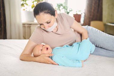 Koruyucu yüz maskesi takmış güzel bir anne kucağında yeni doğmuş oğlunu virüslerden ve enfeksiyonlardan koruyor. Coronavirus covid-19 önleme. Ev karantinası. Evde mutlu bir aile