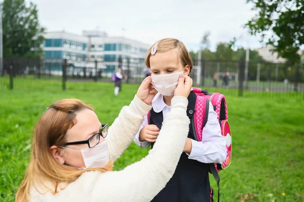 Mother puts a safety mask on her daughter\'s face. Schoolgirls ready go to school. Little sisters with a backpack outdoors. Back to school. Medical mask to prevent coronavirus. Social distance