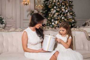 Mutlu Noeller ve mutlu tatiller. Neşeli anne ve tatlı kızı, beyaz klasik iç mekanda bir piyanonun arka planına ve süslü bir Noel ağacına karşı hediyelerini değiş tokuş ediyorlar. Yeni Yıl 2021 