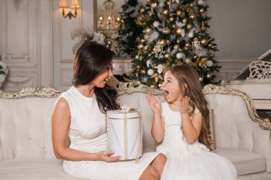 Mutlu Noeller ve mutlu tatiller. Neşeli anne ve tatlı kızı, beyaz klasik iç mekanda bir piyanonun arka planına ve süslü bir Noel ağacına karşı hediyelerini değiş tokuş ediyorlar. Yeni Yıl 2021 