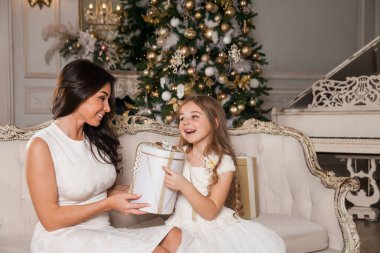 Mutlu Noeller ve mutlu tatiller. Neşeli anne ve tatlı kızı, beyaz klasik iç mekanda bir piyanonun arka planına ve süslü bir Noel ağacına karşı hediyelerini değiş tokuş ediyorlar. Yeni Yıl 2021 