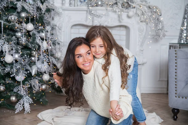 Happy family on New Year Eve. Young beautiful mother and little cute daughter in white cozy sweaters and blanket have fun and hug under a decorated Christmas tree. Festive home classic interior.