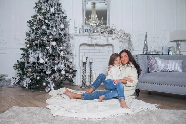 Happy family on New Year Eve. Beautiful mother and cute daughter in white cozy sweaters have fun and hug under a decorated Christmas tree. Festive home classic interior with fireplace and grey sofa.