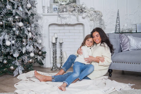 Happy family on New Year Eve. Beautiful mother and cute daughter in white cozy sweaters have fun and hug under a decorated Christmas tree. Festive home classic interior with fireplace and grey sofa.