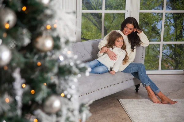 Happy family on New Year Eve. Young beautiful mother and little cute daughter in white cozy sweaters hugging on sofa next to the window and decorated Christmas tree. Festive home classic interior.