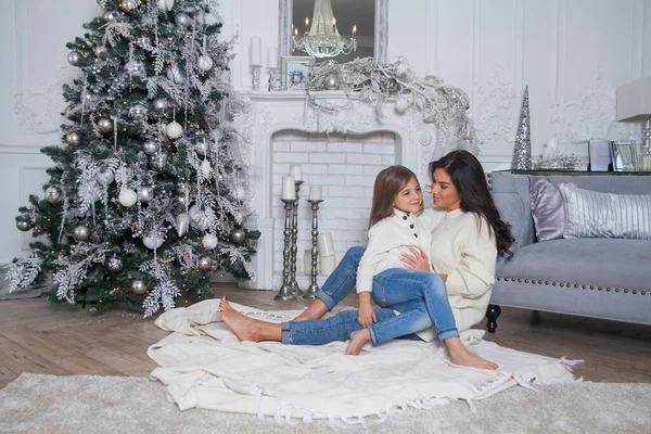 Happy family on New Year Eve. Beautiful mother and cute daughter in white cozy sweaters have fun and hug under a decorated Christmas tree. Festive home classic interior with fireplace and grey sofa.