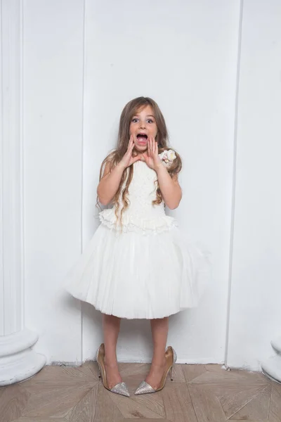 Little princess - excited emotional cute girl in fashion white dress having fun and wearing big mothers sparkle high heels shoes on white background. Free space for text mockup