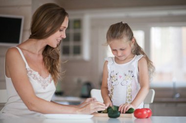 Evde poz veren mutlu bir anne ve çocuk. Güzel bir anne ve küçük kız İskandinav tarzı bir iç mekanda, beyaz bir mutfakta salata için sebze hazırlıyor. Sağlıklı yemek.