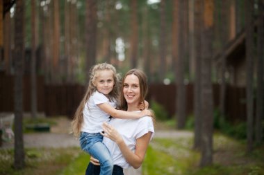 Mutlu gülümseyen annesi ve beyaz tişörtlü ve kot pantolonlu sevimli duygusal kızı kırsal alanda kucaklaşıp eğleniyorlar. Yazı ailemle geçiriyorum.
