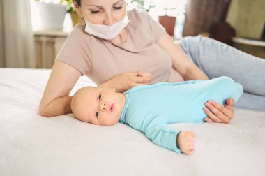 Koruyucu yüz maskesi takmış güzel bir anne kucağında yeni doğmuş oğlunu virüslerden ve enfeksiyonlardan koruyor. Coronavirus covid-19 önleme. Ev karantinası. Evde mutlu bir aile