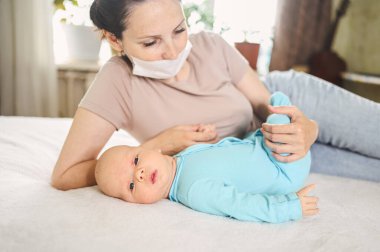 Koruyucu yüz maskesi takmış güzel bir anne kucağında yeni doğmuş oğlunu virüslerden ve enfeksiyonlardan koruyor. Coronavirus covid-19 önleme. Ev karantinası. Evde mutlu bir aile