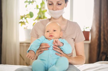 Koruyucu yüz maskesi takmış güzel bir anne kucağında yeni doğmuş oğlunu virüslerden ve enfeksiyonlardan koruyor. Coronavirus covid-19 önleme. Ev karantinası. Evde mutlu bir aile