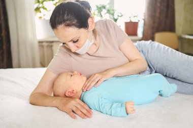 Koruyucu yüz maskesi takmış güzel bir anne kucağında yeni doğmuş oğlunu virüslerden ve enfeksiyonlardan koruyor. Coronavirus covid-19 önleme. Ev karantinası. Evde mutlu bir aile