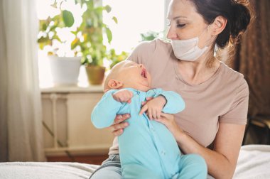 Koruyucu yüz maskesi takmış güzel bir anne kucağında yeni doğmuş oğlunu virüslerden ve enfeksiyonlardan koruyor. Coronavirus covid-19 önleme. Ev karantinası. Evde mutlu bir aile