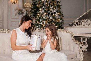 Mutlu Noeller ve mutlu tatiller. Neşeli anne ve tatlı kızı, beyaz klasik iç mekanda bir piyanonun arka planına ve süslü bir Noel ağacına karşı hediyelerini değiş tokuş ediyorlar. Yeni Yıl 2021 