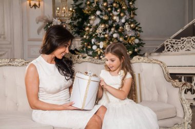 Mutlu Noeller ve mutlu tatiller. Neşeli anne ve tatlı kızı, beyaz klasik iç mekanda bir piyanonun arka planına ve süslü bir Noel ağacına karşı hediyelerini değiş tokuş ediyorlar. Yeni Yıl 2021 