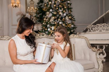 Mutlu Noeller ve mutlu tatiller. Neşeli anne ve tatlı kızı, beyaz klasik iç mekanda bir piyanonun arka planına ve süslü bir Noel ağacına karşı hediyelerini değiş tokuş ediyorlar. Yeni Yıl 2021 