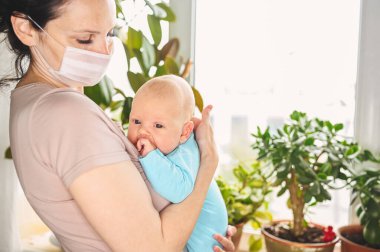Koruyucu yüz maskesi takmış güzel bir anne kucağında yeni doğmuş oğlunu virüslerden ve enfeksiyonlardan koruyor. Coronavirus covid-19 önleme. Ev karantinası. Ailesi pencerenin yanında.