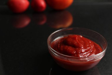 Kırmızı domates soslu bir cam tencere arka planda sebzelerin yanında siyah bir zemin üzerinde duruyor. Kırmızı domates, turuncu domates ve sarı kabuklu soğan.