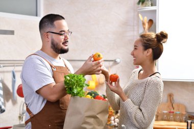 Genç Asyalı çifti sevmek mutfakta yemek yapmak sağlıklı yemekler yapmak ve sebzelerle alışveriş torbalarını birlikte tutmak eğlenceli geliyor.