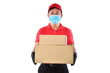 Kırmızı üniformalı, tıbbi maskeli, koruyucu eldivenli mutlu Asyalı teslimatçı beyaz arka planda karton kutu taşıyor. Teslimatçı çocuk paket teslimatı yapıyor. Güvenli teslimat.
