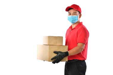 Kırmızı üniformalı, tıbbi maskeli, koruyucu eldivenli mutlu Asyalı teslimatçı beyaz arka planda karton kutu taşıyor. Teslimatçı çocuk paket teslimatı yapıyor. Güvenli teslimat.           